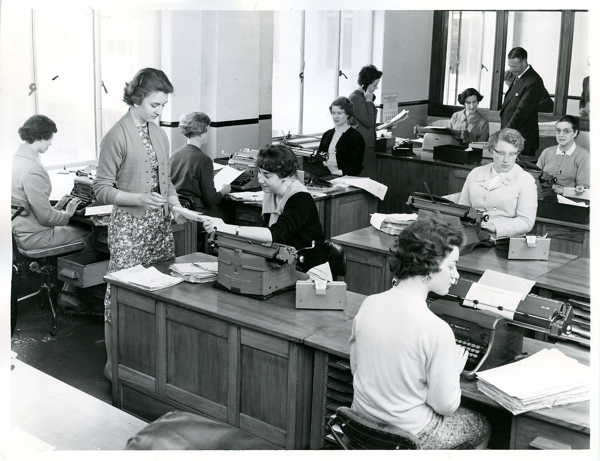 A 1959 office: rows of typists at desks with typewriters and stacks of paper, a supervisor handing over a document.